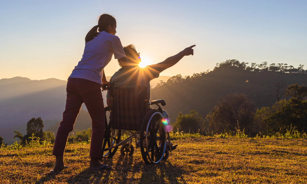 A woman with a ponytail and a man who is a wheelchair user in front of a glowing sunrise and background of mountains and trees. The man is pointing to something out of frame, and the image conveys travel and adventure.