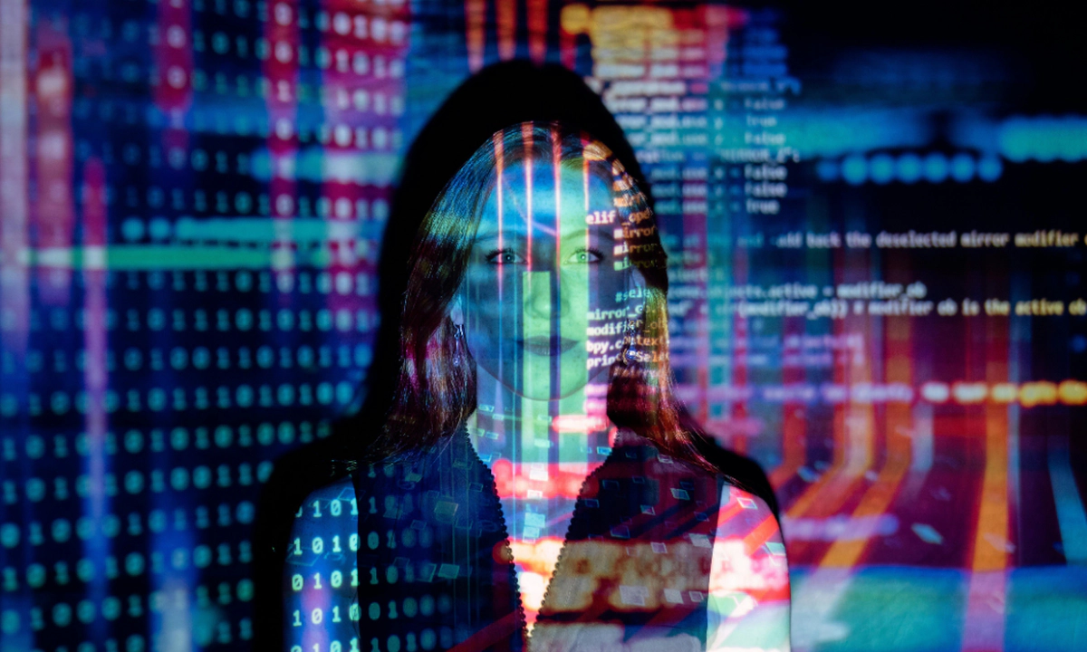 Photo of a woman standing in front of a wall facing the camera with colourful lights, lines of code, text and shapes projected onto her to convey tech.