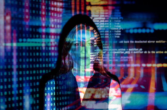 Photo of a woman standing in front of a wall facing the camera with colourful lights, lines of code, text and shapes projected onto her to convey tech.