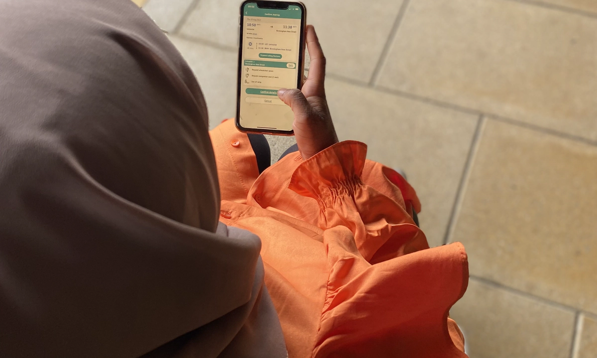 woman in a brown headscarf and orange long sleeved dress holding a smartphone. The woman is a wheelchair user and she is on a train platform. Her phone screen shows the Passenger Assistance app.