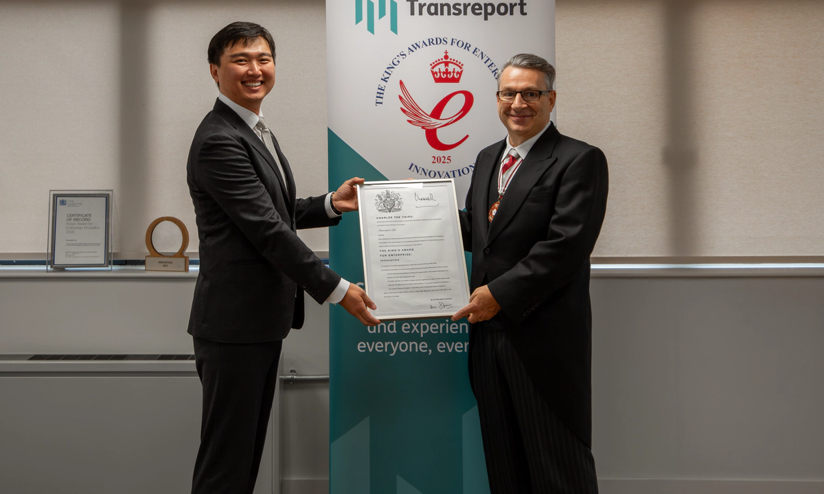 Photo of Jay Shen and Mr Kevin McGrath OBE MRICS DL smiling, holding the official King’s Award certificate together. They stand in front of the Transreport King’s Award for Enterprise in Innovation banner.