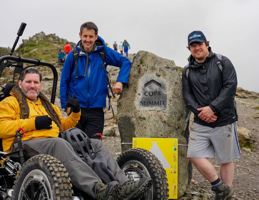 Nick Wilson in RockClimber beside the Copa Summit sign on Mount Snowdon. Nick is smiling, wearing a yellow hoodie, grey trousers, and black gloves. Nick Wilson in RockClimber beside the Copa Summit sign on Mount Snowdon. Nick is smiling, wearing a yellow hoodie, grey trousers, and black gloves.