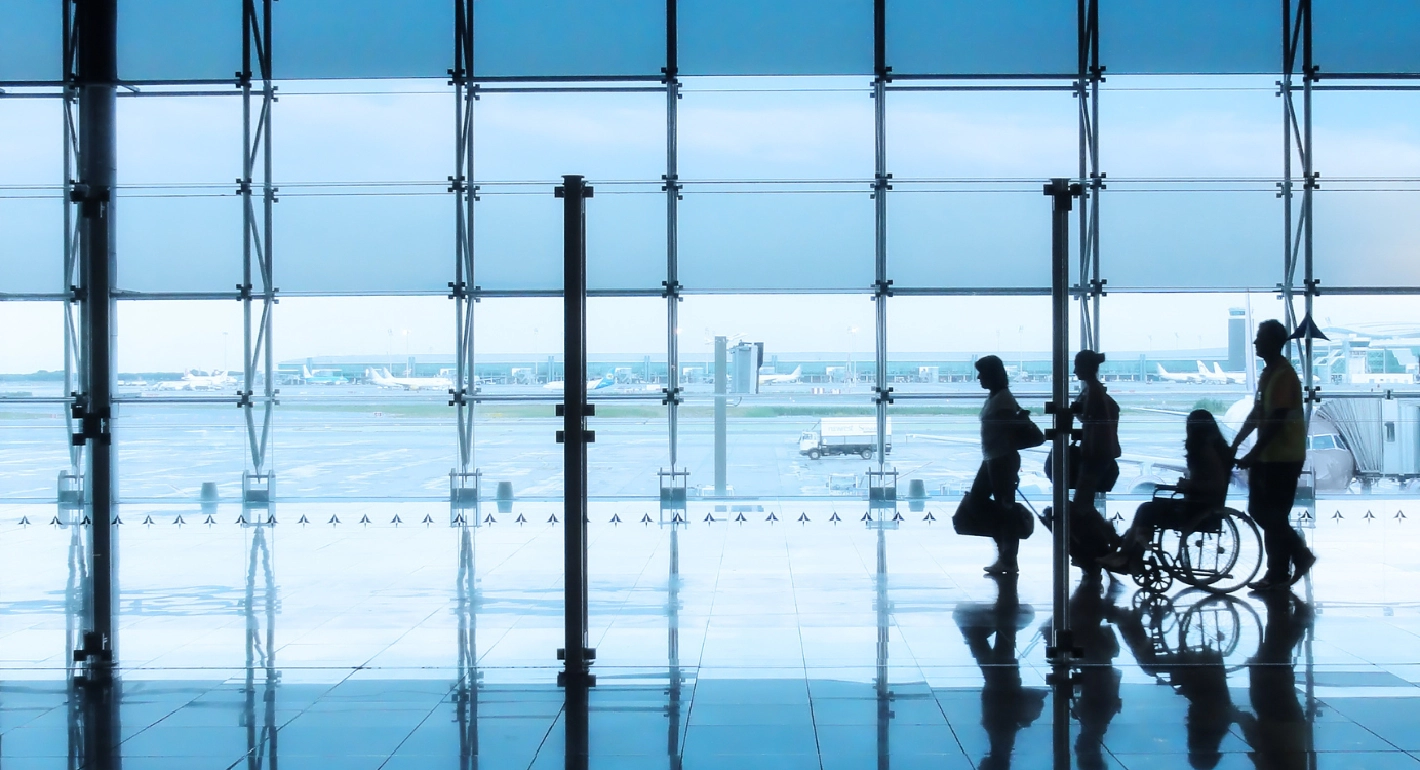 Silhouettes of four people, three walking and one in a wheelchair, in an airport lounge with an aeroplane runway in the background.