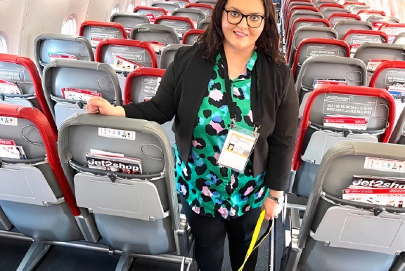 Emma Partlow, Transreport's Inclusion & Accessibility Manager, standing beside her assistance dog Luna, in the middle aisle of a commercial passenger aeroplane.