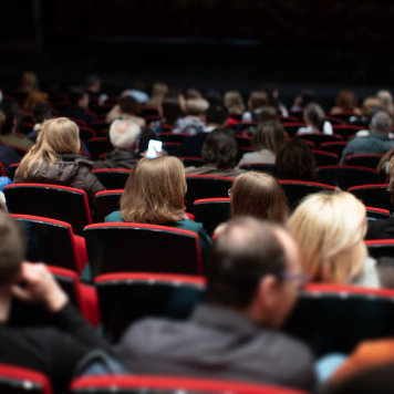 A photo of people at a venue seated in red chairs in the audience. A photo of people at a venue seated in red chairs in the audience.