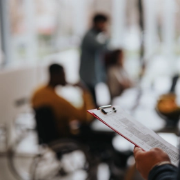 Restaurant Photo with a blur effect of a restaurant. The person in focus holds a clipboard with information. In the background are three people including a man in a yellow jumper who is a wheelchair user.