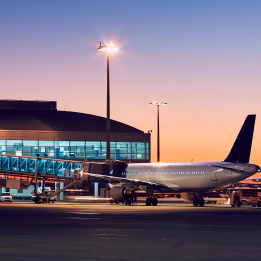 A stationary commercial passenger aeroplane standing on a runaway, with an airport terminal in the background against a sunset.