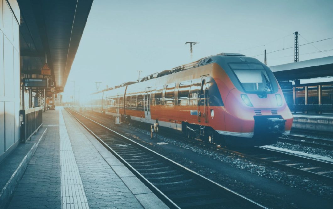 A red and black commercial passenger train, stood stationary at a train platform, overlayed with a white version of the Transreport logo.