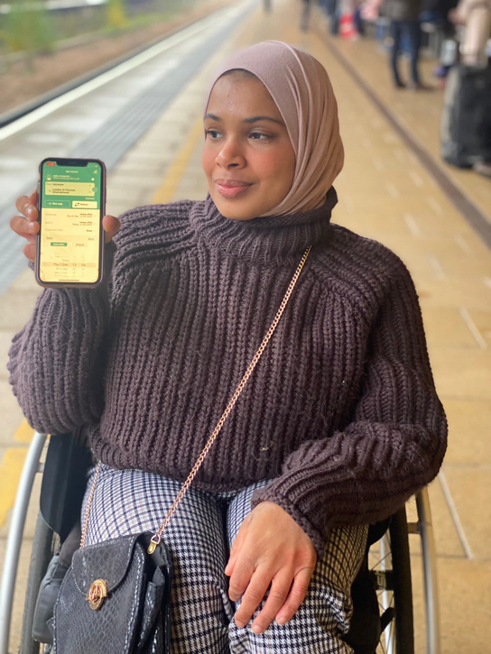 Umaymah Dakri, a woman with a brown jumper, headscarf and checked trousers. Umaymah is a wheelchair user and holds a smart phone displaying the New Journey page in the PA App.
