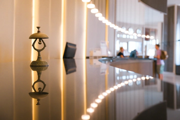 Photo of a hotel check-in desk with a bell in the foreground and a customer speaking to a member of staff in the background.