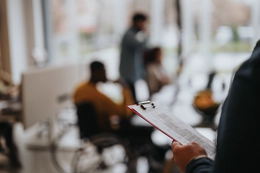Photo with a blur effect of a restaurant. The person in focus holds a clipboard with information. In the background are three people including a man in a yellow jumper who is a wheelchair user.
