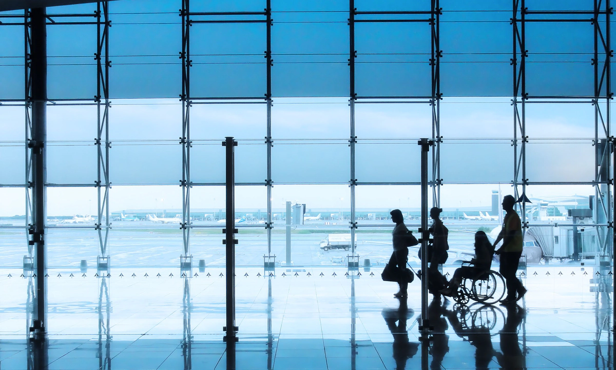 Four silhouettes of people in an airport setting, with a man and woman walking together with suitcases, followed by a wheelchair user being pushed by another person