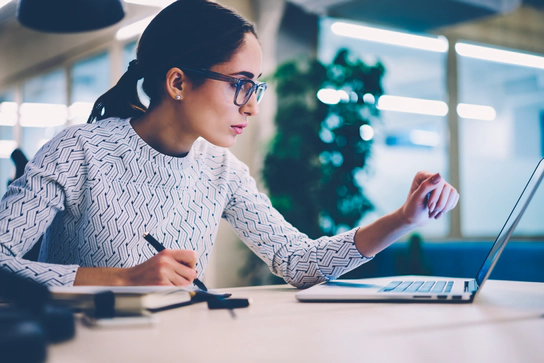 A woman with a ponytail and glasses taking notes as she looks at a computer screen.