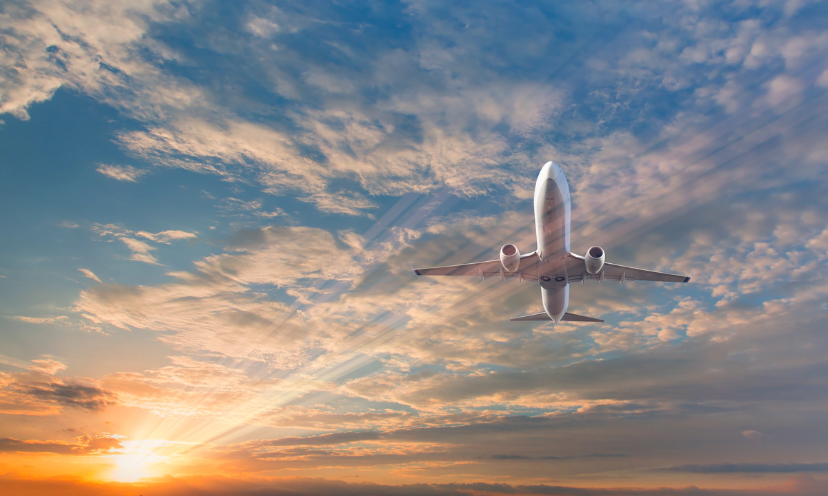 A picture of a white passenger plane from below, against a blue sky filled with clouds and a lens flare from the sun