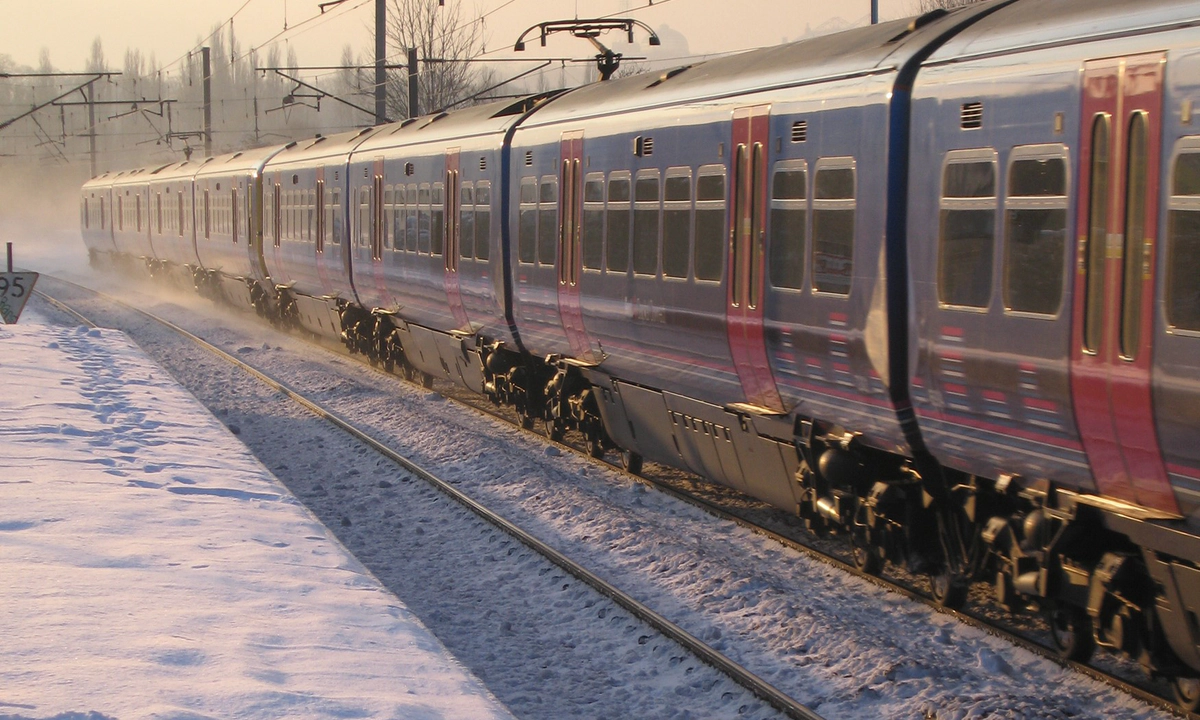 Photo of a blue and red train on a snowy track beside a snowy platform with an orange sky behind it.