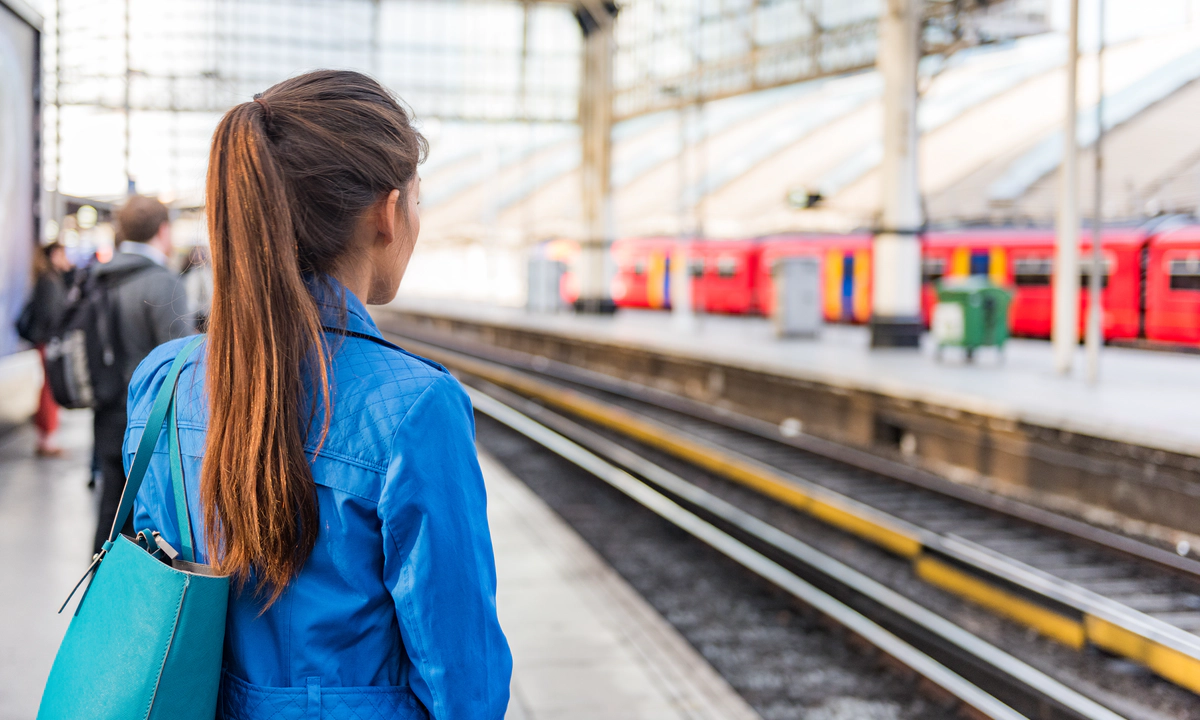 Photo of a train platform. The platform and people on the platform are blurred except for a woman in the foreground who stands with her back to the camera with long brown hair in a ponytail and a blue denim jacket. A red train is waiting at the platform opposite.