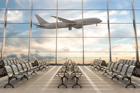 An empty airport lounge with floor to ceiling glass windows looking out onto an airport runway, with a commercial passenger aeroplane mid-take off in the background.