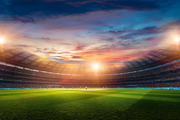 A photo of a sports stadium with bright green grass, players on the pitch, and an open sky above.
