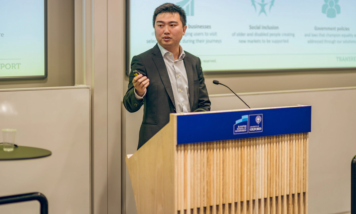 Jay Shen, an asian man with short black hair, and CEO of Transreport, in a grey suit standing at a speaking podium.