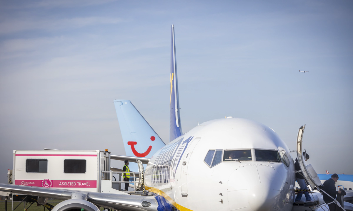 A zoomed in photograph of a Ryainair Boeing 747 plane from the front, with passengers disembarking by the front exit, by stairs, on the right, and an ambulift docking with the central exit of the plane on the left.