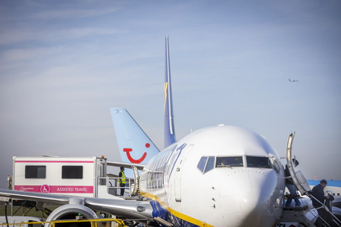 A zoomed in photograph of a Ryainair Boeing 747 plane from the front, with passengers disembarking by the front exit, by stairs, on the right, and an ambulift docking with the central exit of the plane on the left.